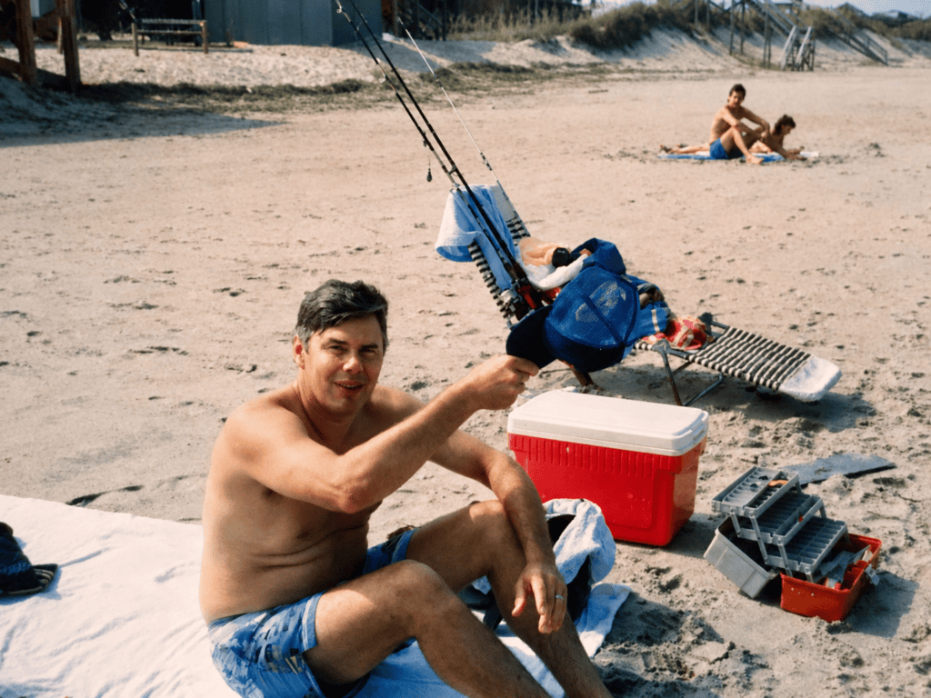 Roger Hamrick sitting on a beach beside fishing rods, a red cooler, and tackle boxes during a family trip.
