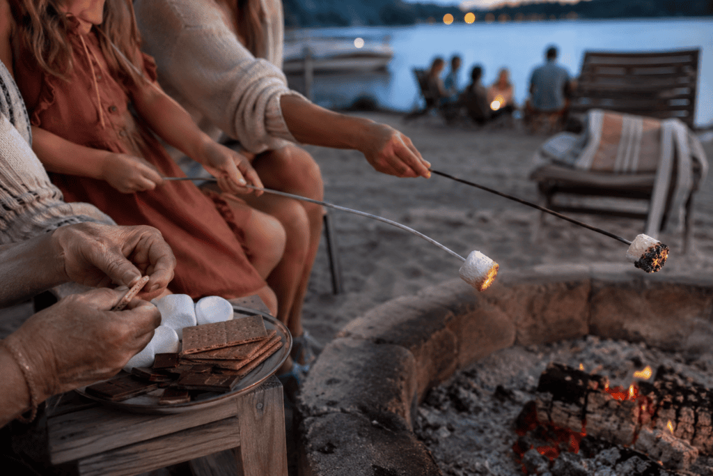 Three generations roasting marshmallows for s’mores beside a lakeside fire pit.