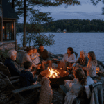 Multigenerational family roasting marshmallows around a lakeside fire pit at dusk.