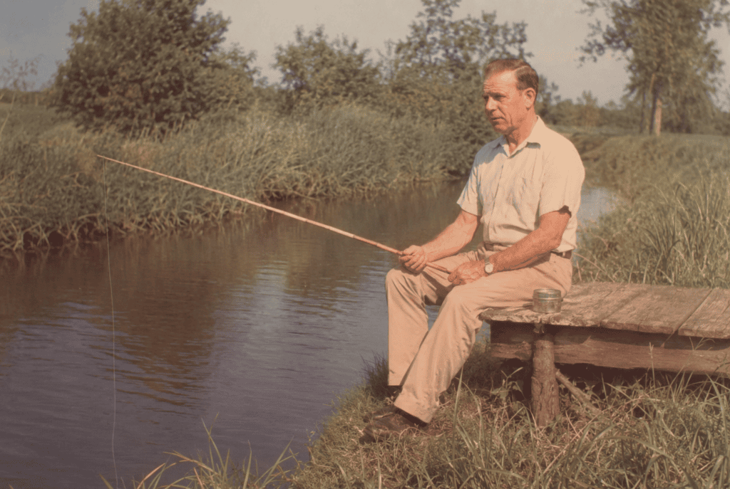 Granddaddy sitting on a short wooden dock with a cane pole extended over the quiet canal water.