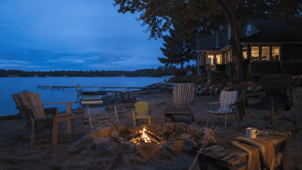 Empty chairs surrounding a fading fire beside a quiet lake house at blue hour.