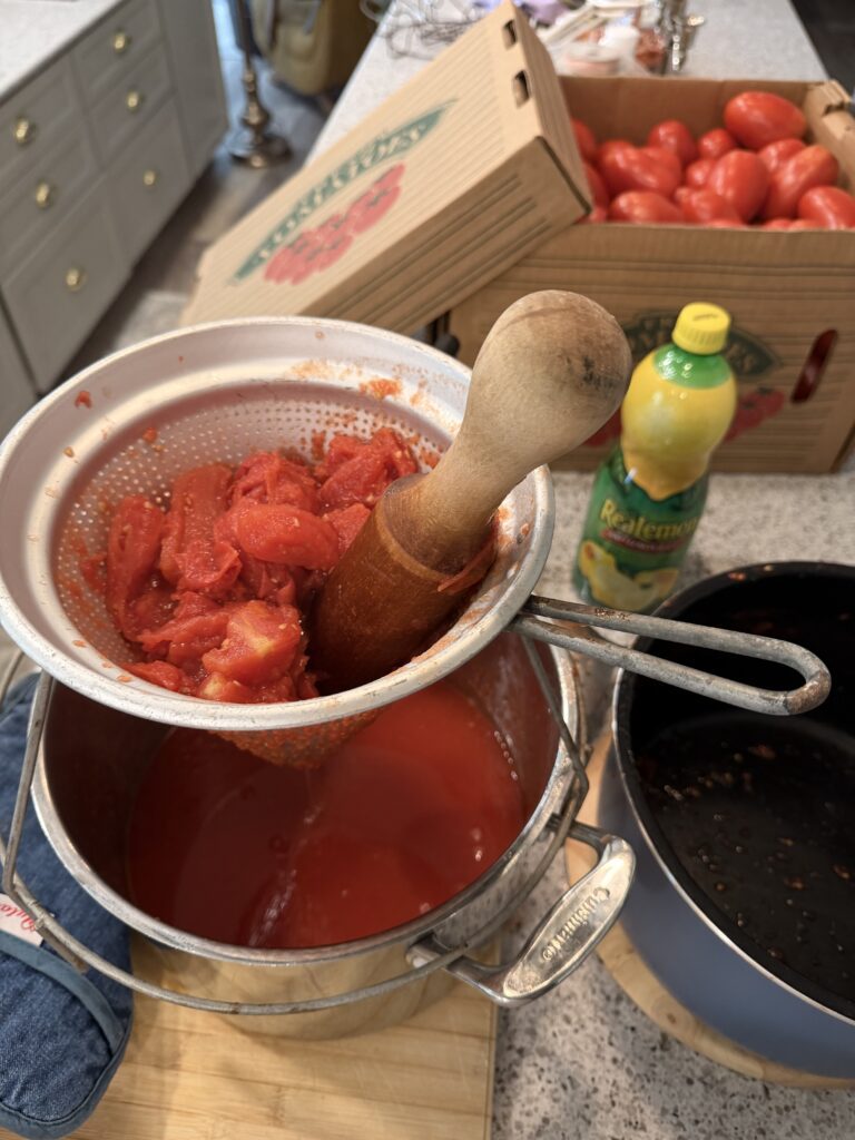 Cooked tomatoes being pressed through a metal sieve with a wooden pestle into a pot of fresh tomato sauce.